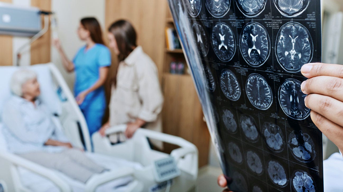 1439950058 MRI of head of elderly woman in hands of doctor standing in medical ward near senior patient with relative and nurse. Recovery after a stroke