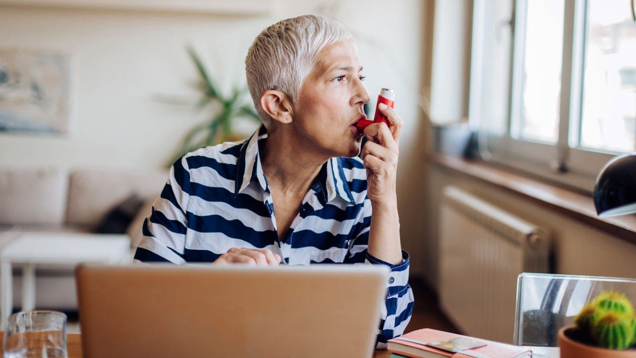 Mature woman using asthma inhaler while working on a laptop at home.