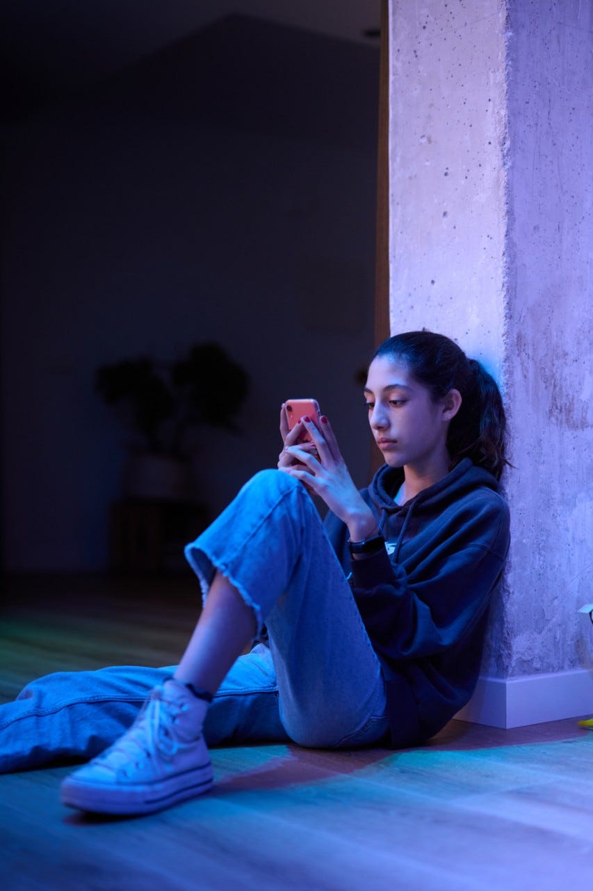 Portrait of a brunette teen girl in jeans and hoodie chatting via cell phone while sitting back at wall at home.