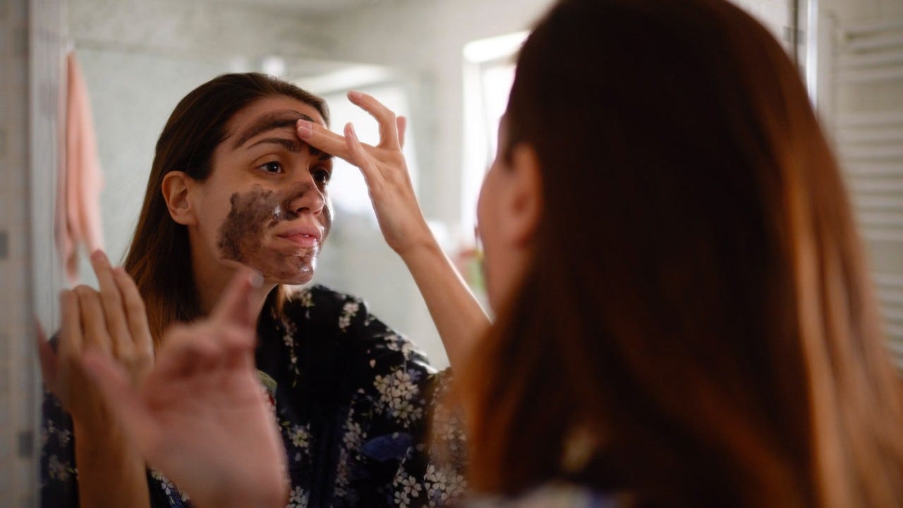 1008336154 Young female in the bathroom looking in the mirror and taking care of her facial skin.