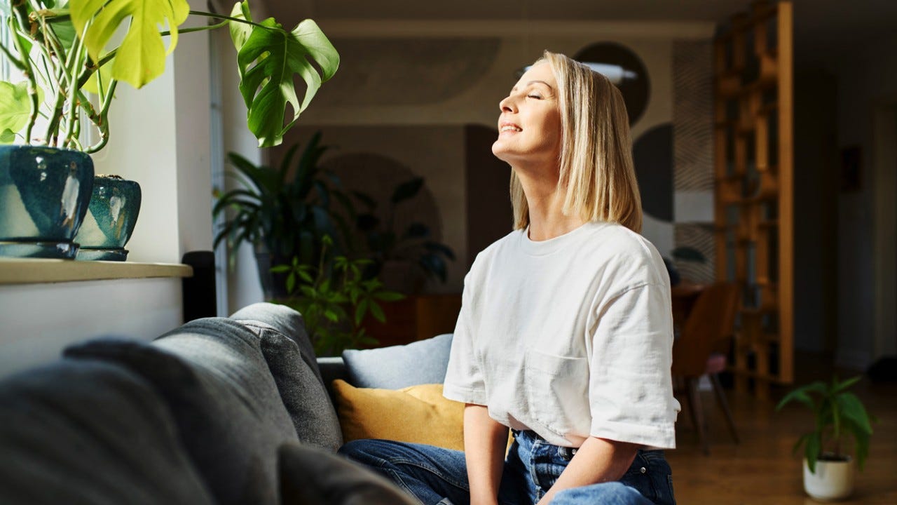 Happy adult woman sitting on the sofa with eyes closed enjoying bright daylight