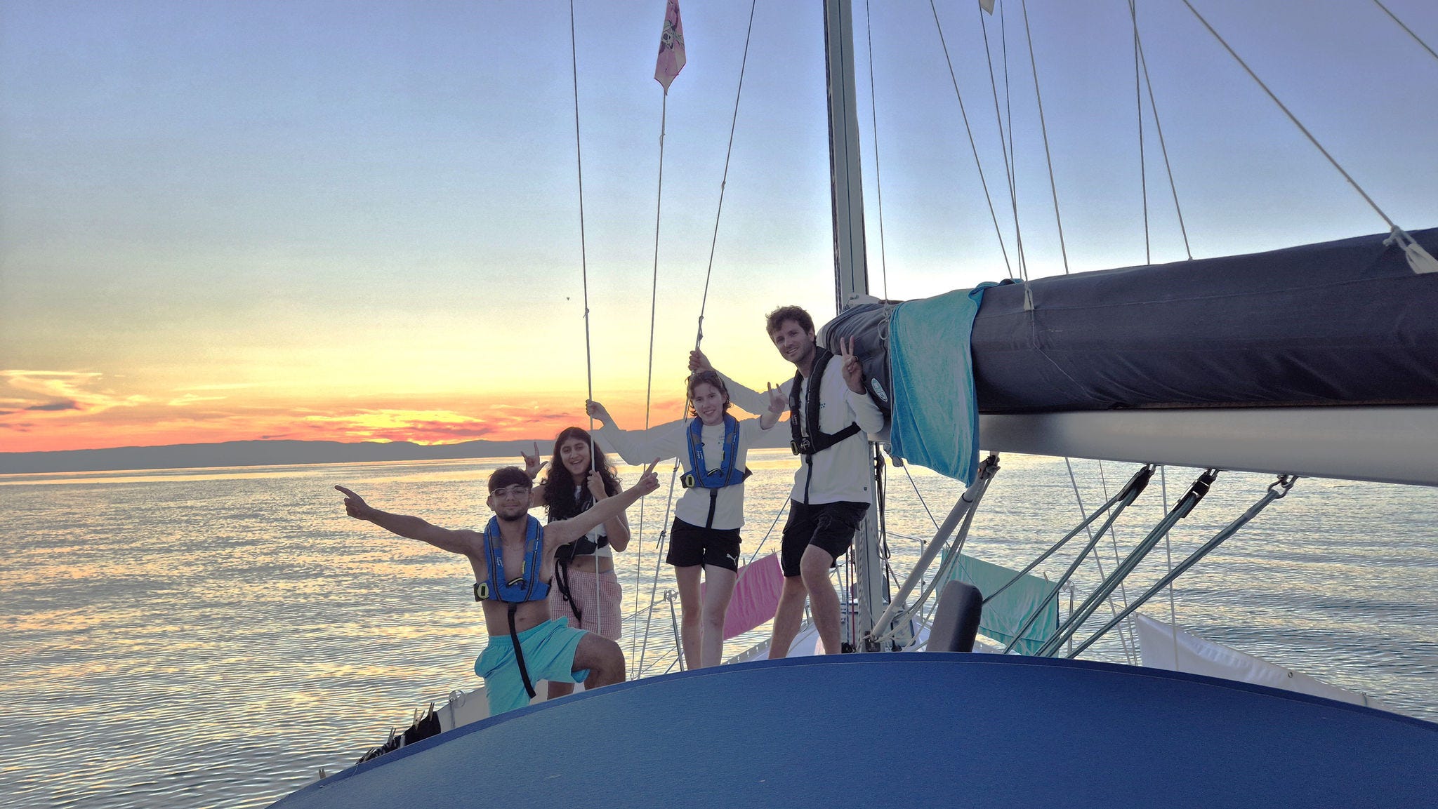 Freunde auf einem Segelboot bei Sonnenuntergang, stehend am Bug über ruhigem Meer, entspannt und fröhlich.