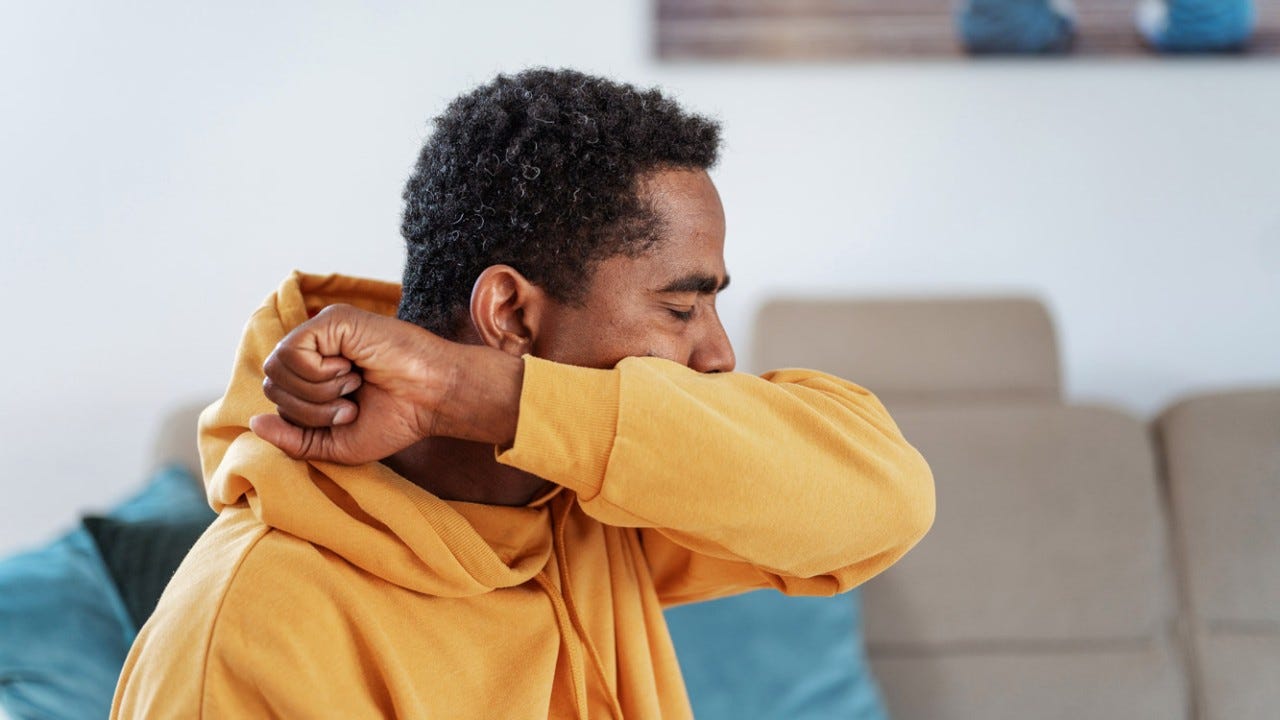 Adult man wearing a yellow hoodie in a living room, coughing or sneezing into elbow.